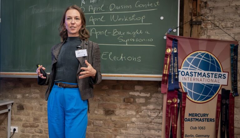 Join Meetings and Inspire others ,Sarah Basteck delivering a speech at Mercury Toastmasters Berlin, standing confidently in front of a chalkboard with a Toastmasters banner beside her.
